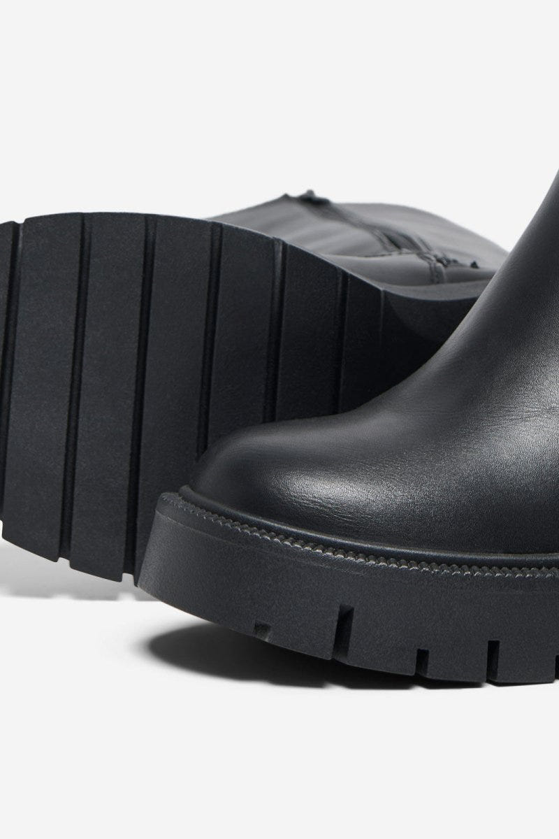 Close-up of black leather boot with chunky lug sole and rounded toe on a light grey background