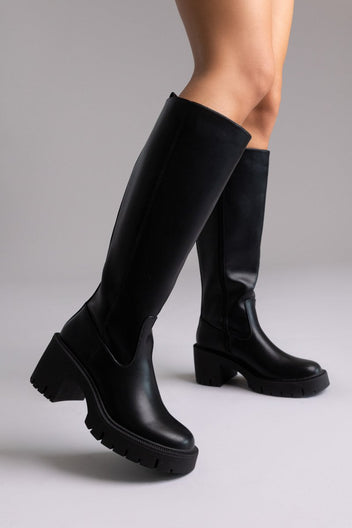 Model leg raised in black leather knee-high boot with chunky block heel and lug sole, angled view on a light background