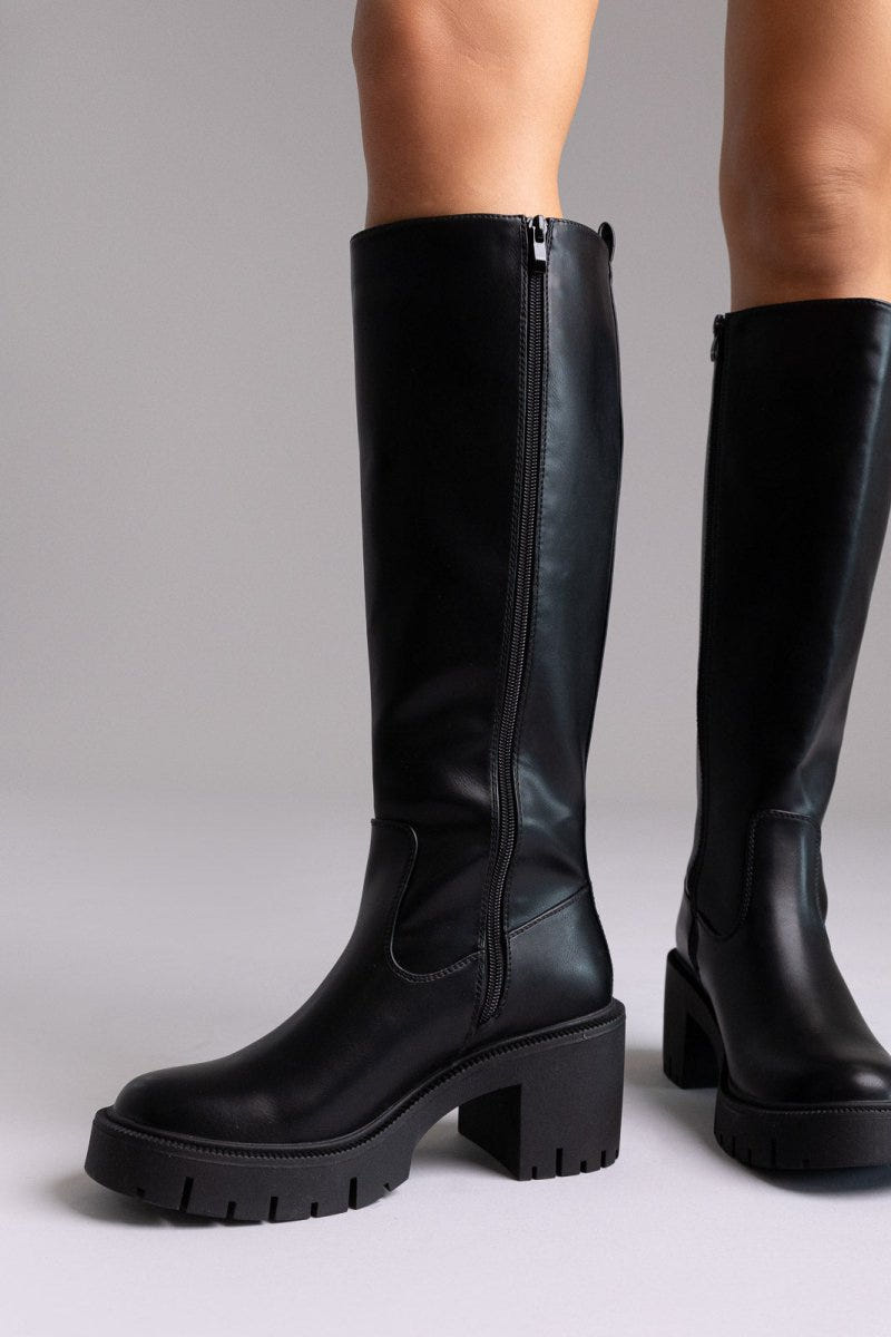 Model legs in black leather knee-high boots with chunky lug sole and side zip, front view on a light lilac background