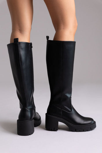 Model legs in black leather knee-high boots with chunky block heels and side zip, front view on a light lilac background