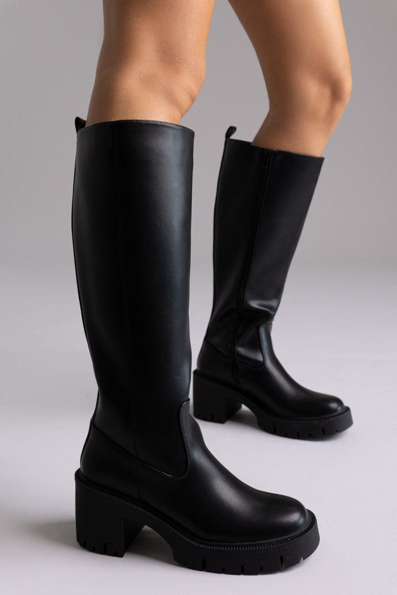 Model legs in black leather knee-high boots with chunky block heels and side zip, angled view on a light lilac background