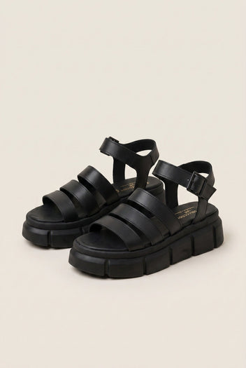Pair of black chunky platform sandals with wide straps and buckle closure, angled view on a light beige background