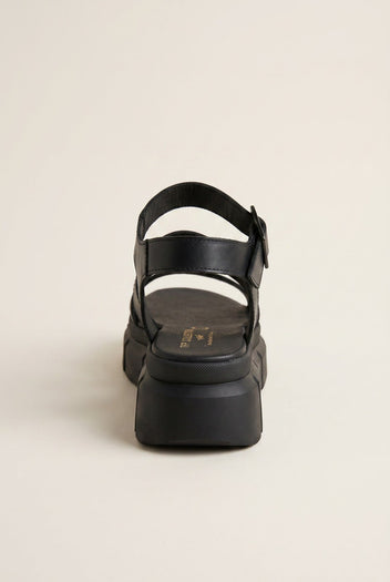 Black chunky platform sandal with ankle strap and buckle, rear view on a light beige background
