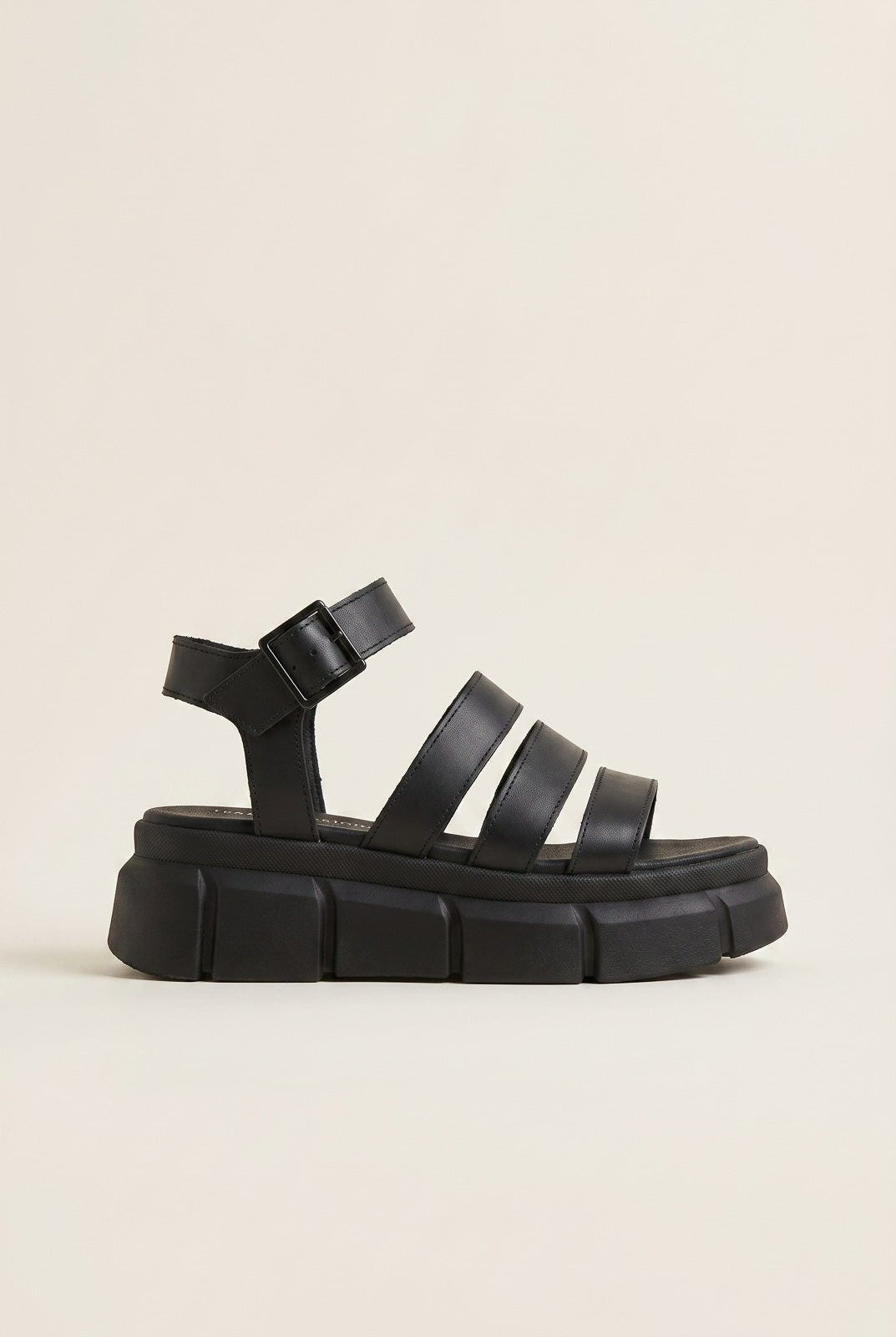 Black chunky platform sandal with wide straps and ankle buckle, side view on a light beige background