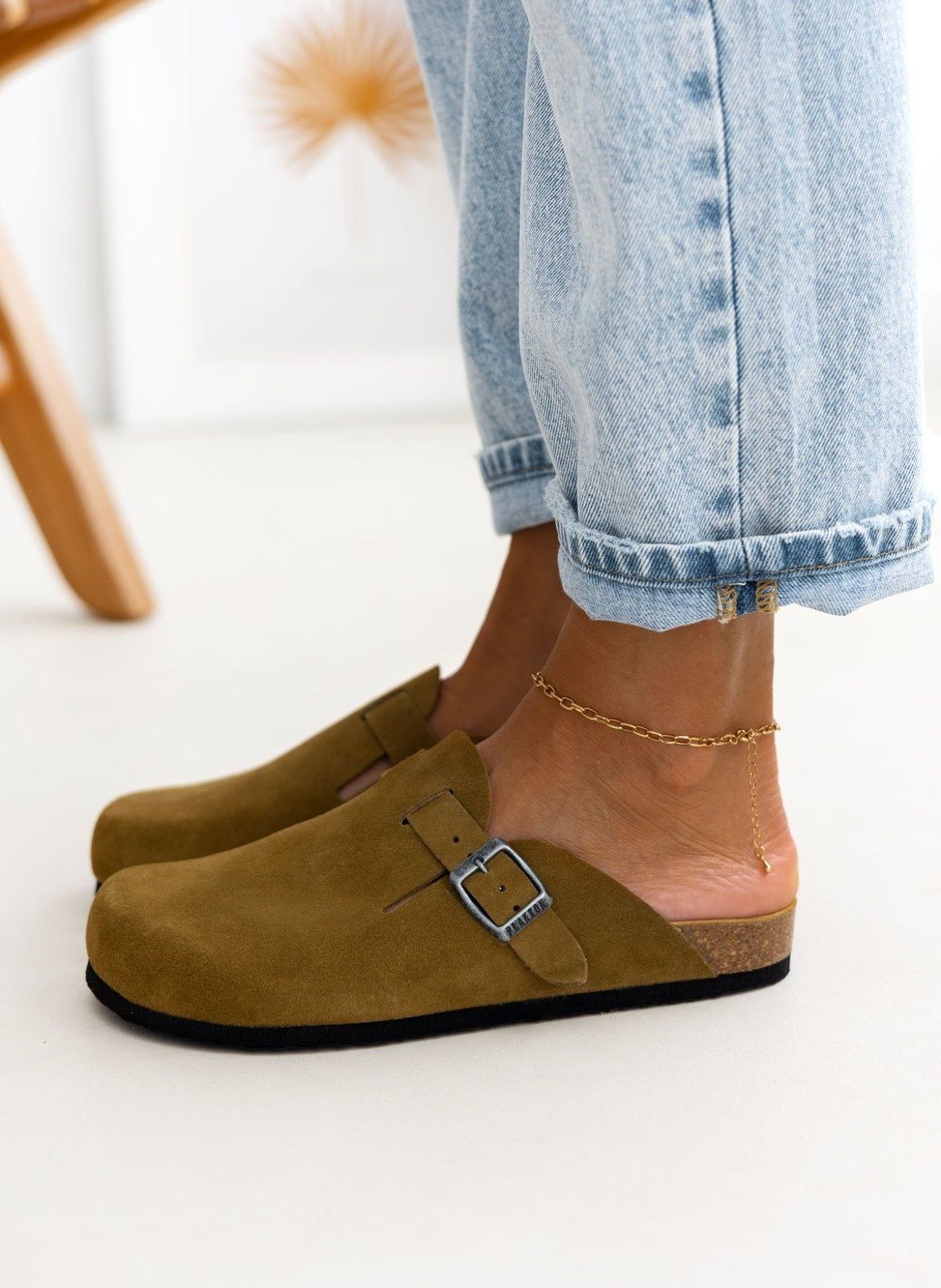 Model foot in olive suede clog with buckle strap and cork footbed in front of rolled light denim jeans