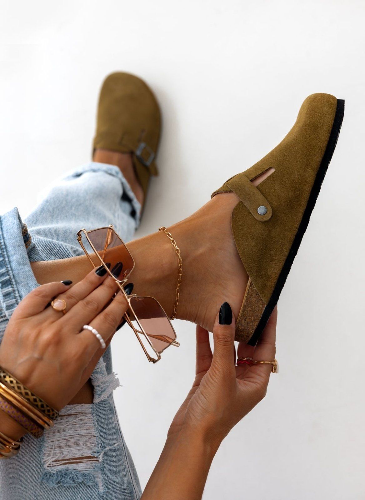 Close-up side view of olive suede clog held in hand near model ankle with bracelets and rolled denim