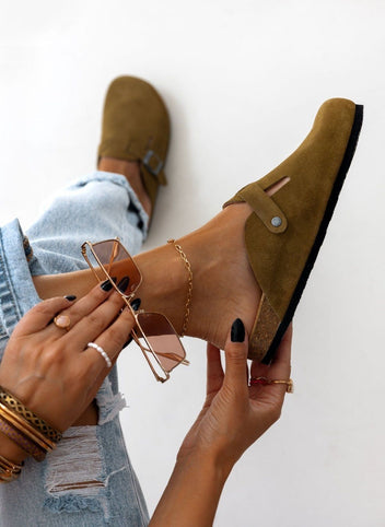 Close-up side view of olive suede clog held in hand near model ankle with bracelets and rolled denim