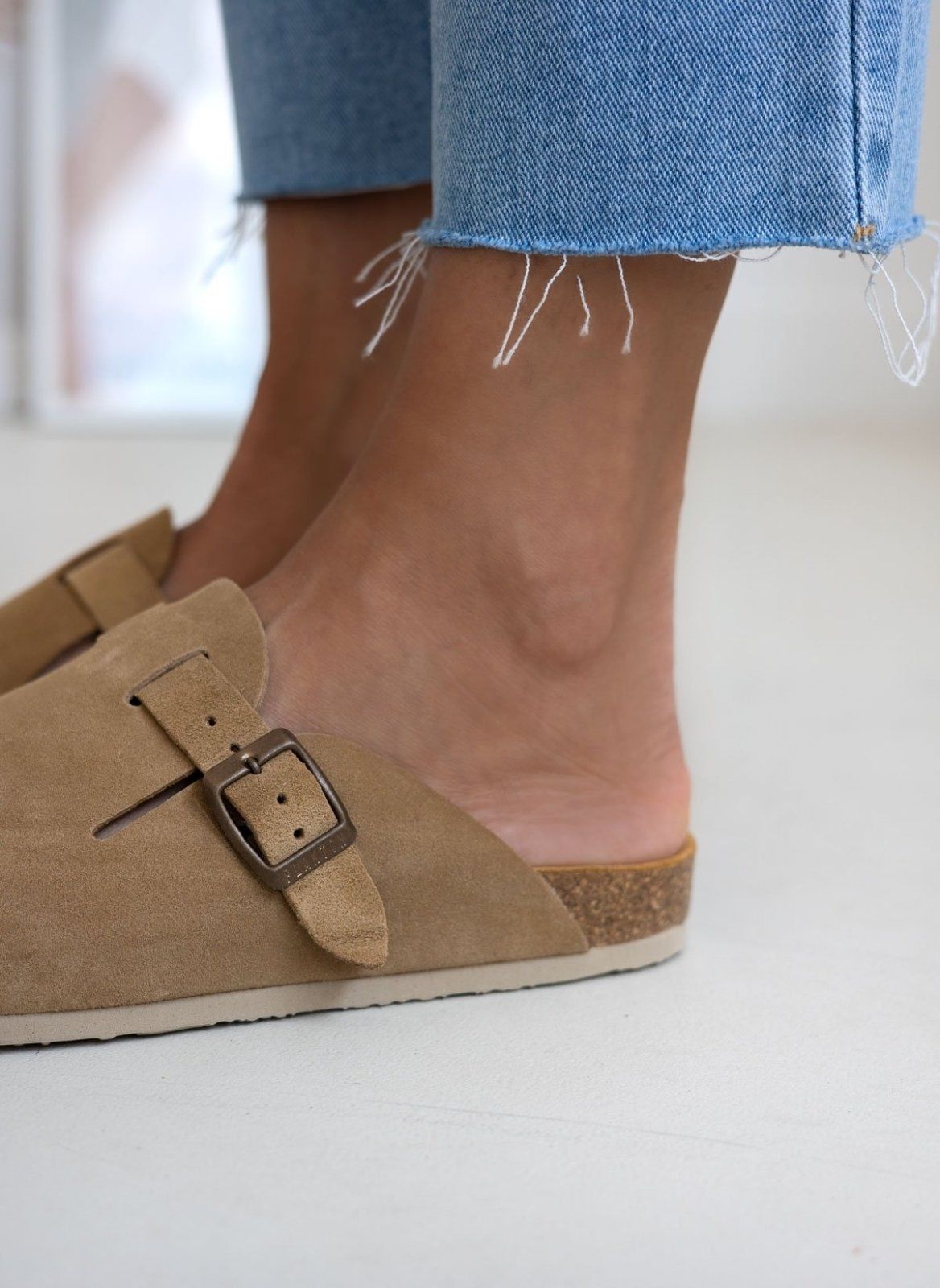 Close-up of beige suede clog buckle strap worn on model ankle with frayed denim hem above