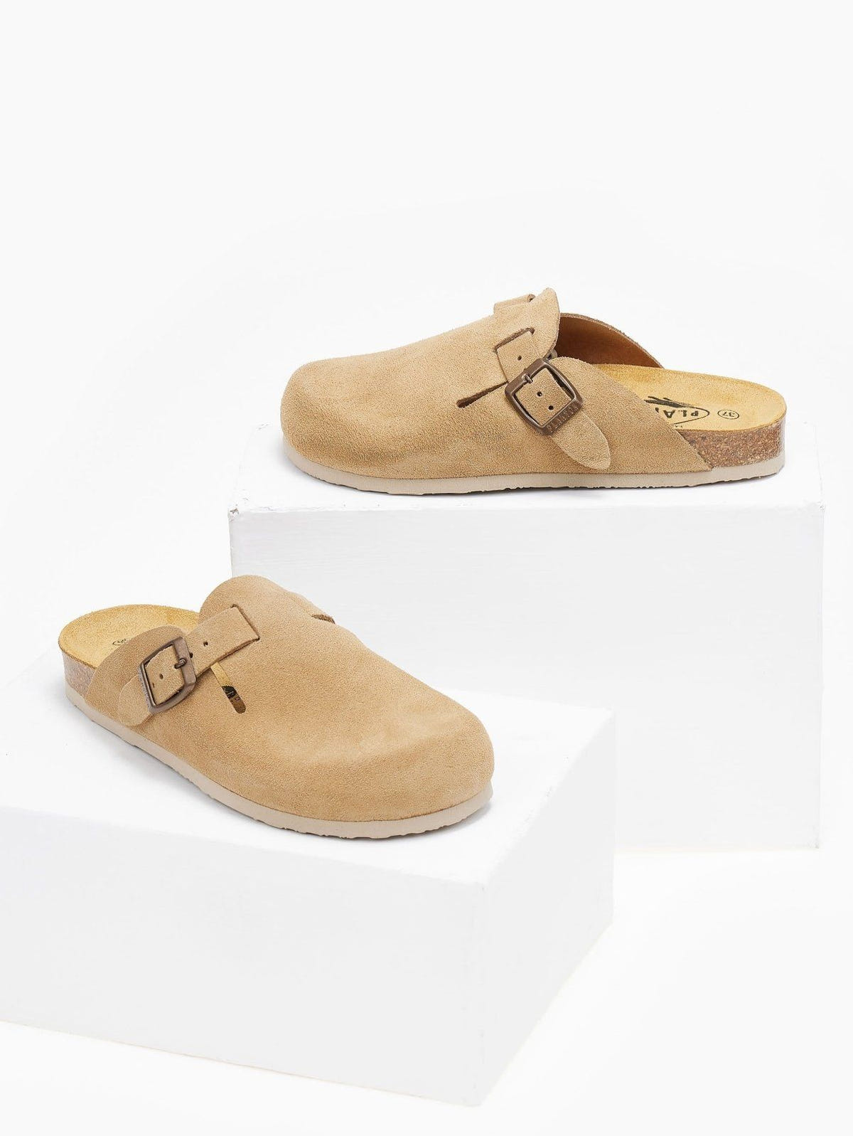 Pair of beige suede clogs on white blocks showing buckle straps and cork footbed from a low angle