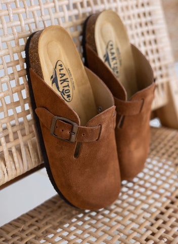 Pair of brown suede clogs with buckle strap, angled view on a woven basket backdrop