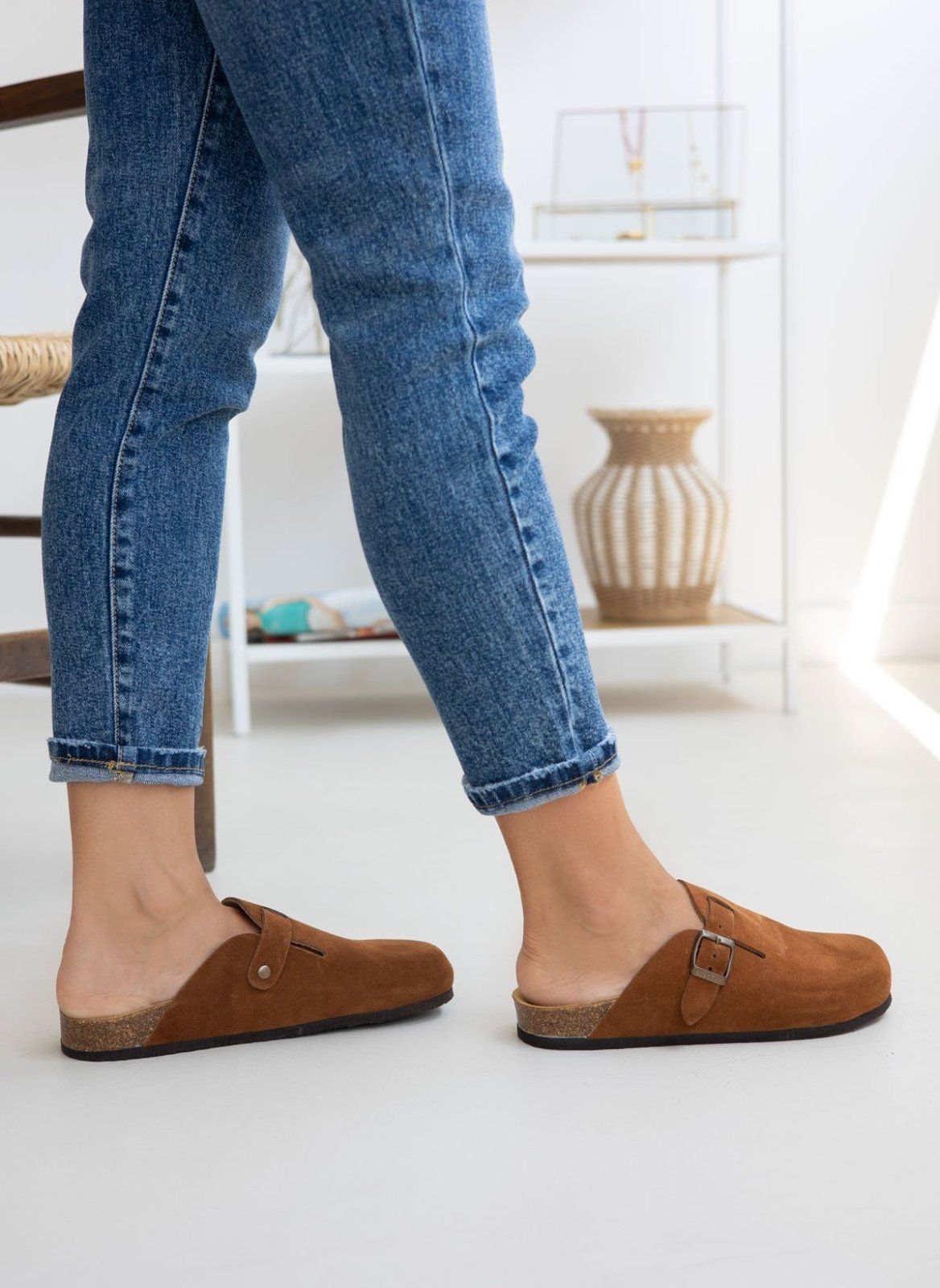 Model legs in brown suede clogs with buckle strap, worn with light blue jeans, angled view on a light background