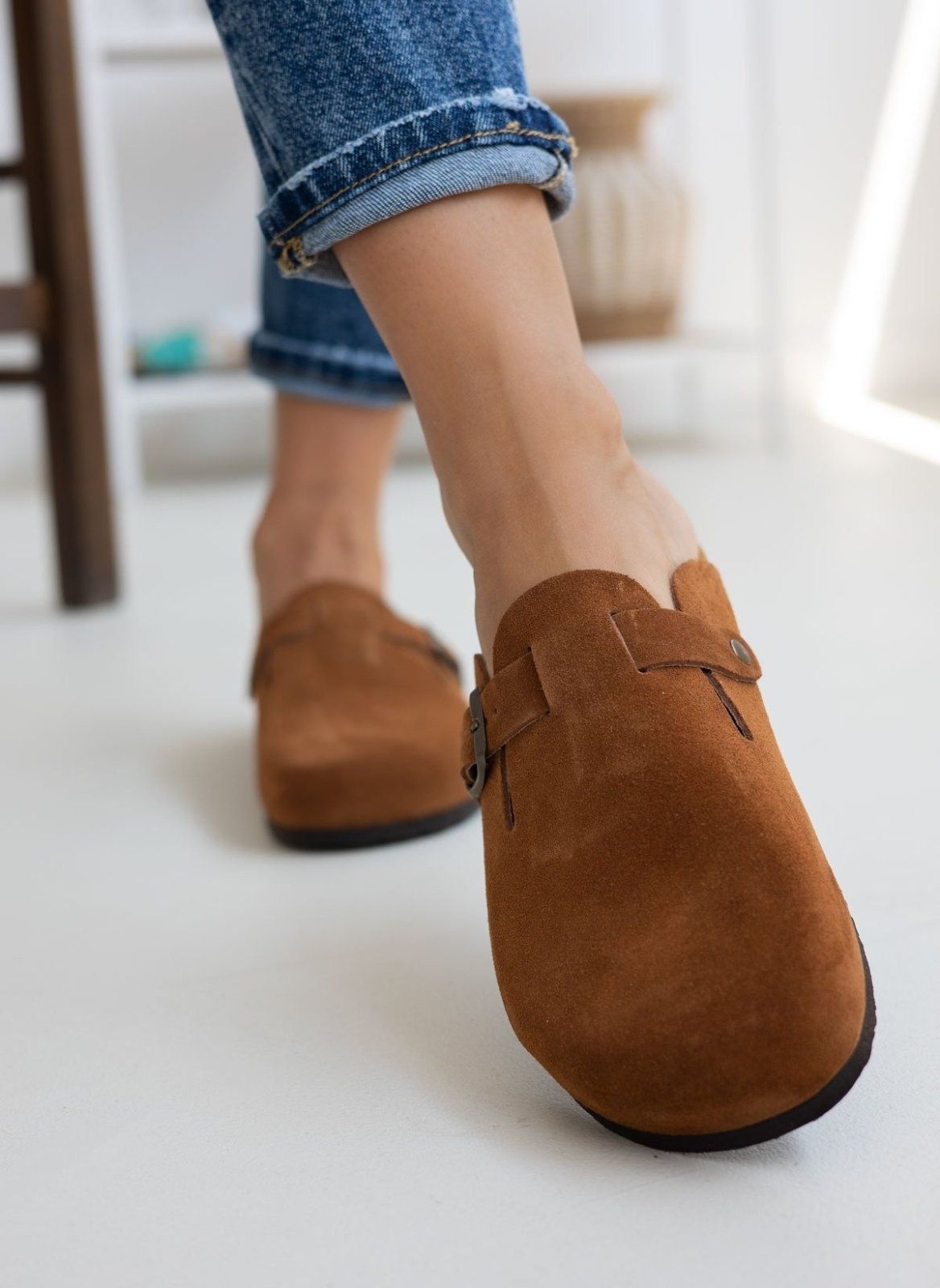 Model foot in brown suede clog with buckle strap, worn with light blue jeans, angled view on a light background