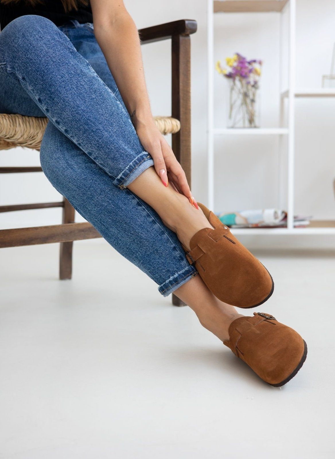 Model legs in brown suede clogs with buckle strap, worn with light blue jeans, seated on a chair, side view