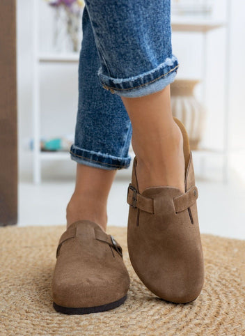 Model legs in taupe suede clogs with buckle strap, worn with light blue cuffed jeans, angled view on a woven rug