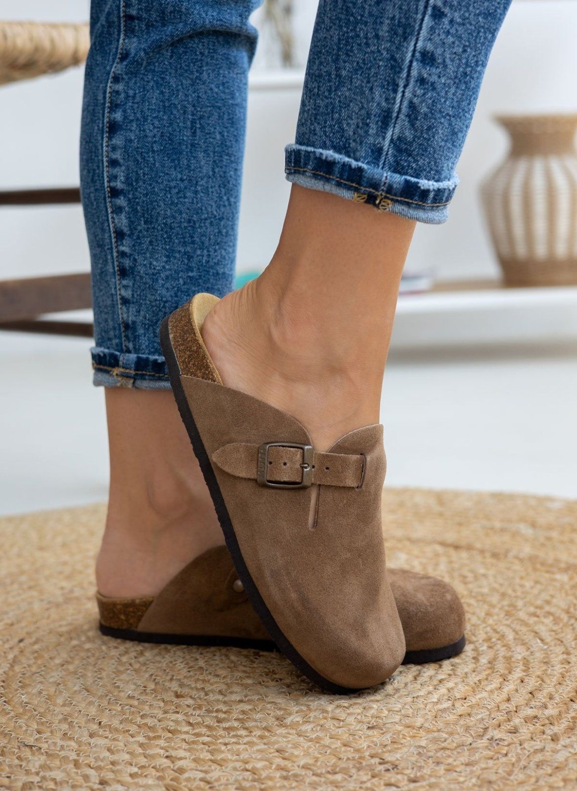 Model foot in taupe suede clog with buckle strap on round woven mat beside rolled denim jeans