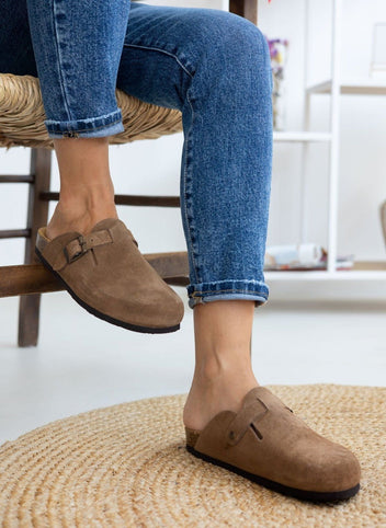 Model legs in taupe suede clogs with buckle strap, worn with light blue jeans, seated on a chair, front view