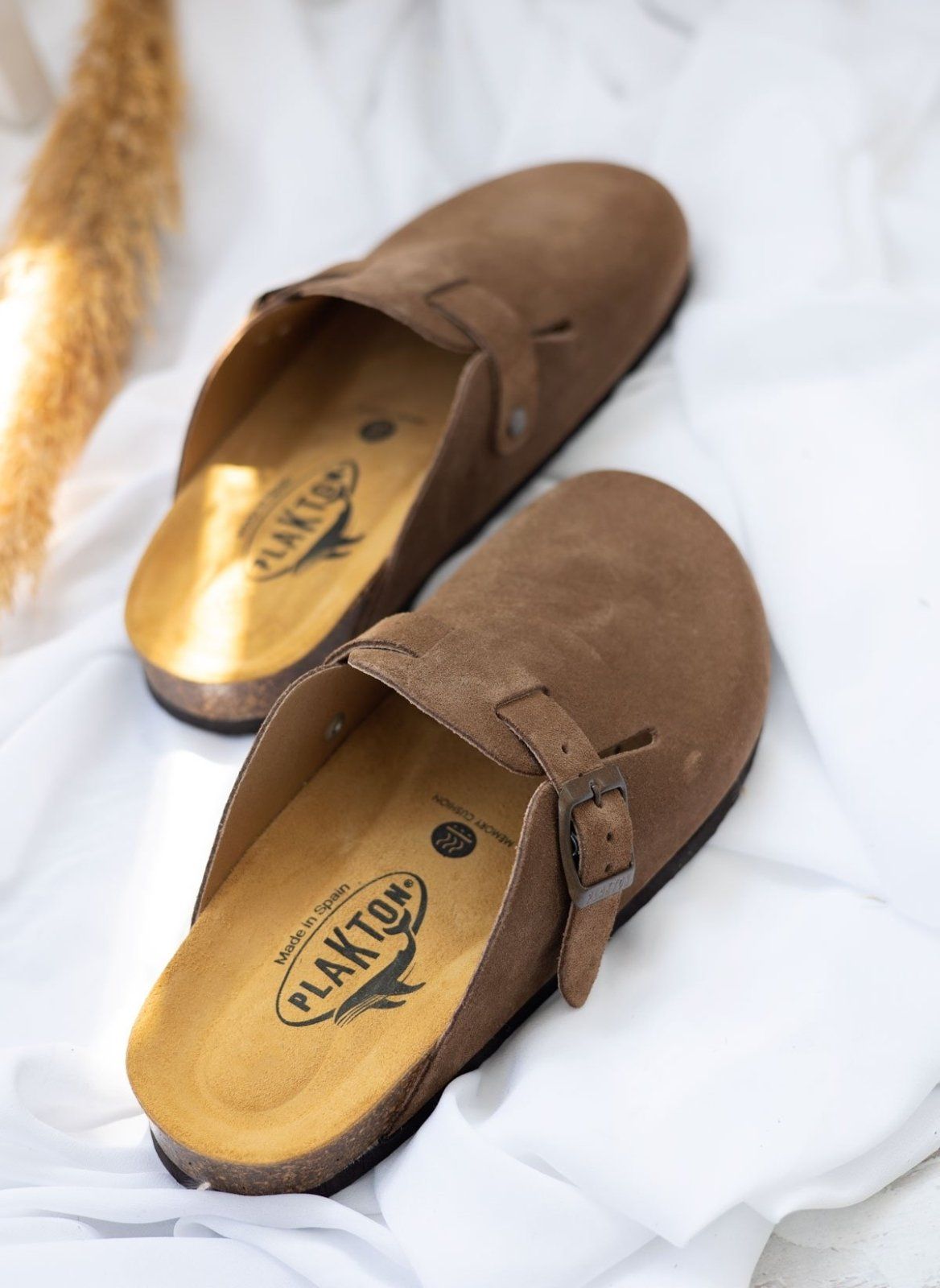 Taupe suede clogs with buckle straps angled to show golden cork footbed resting on white fabric