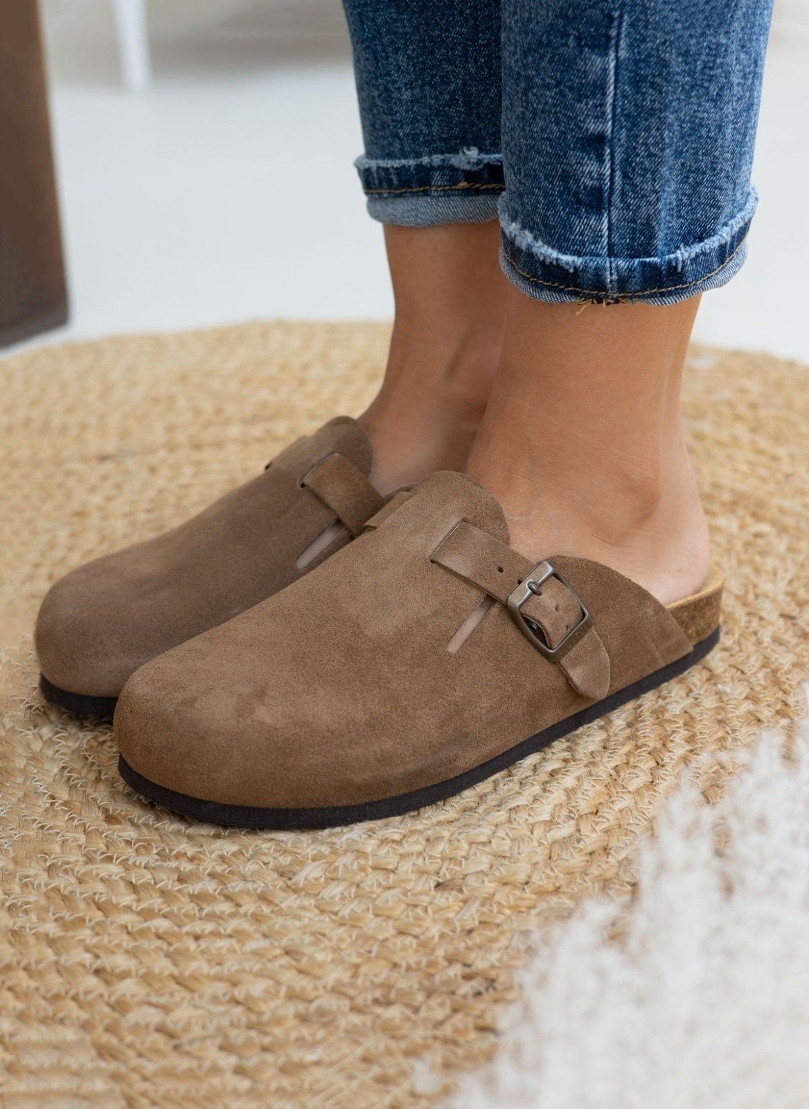 Model foot in taupe suede clog with buckle strap, worn with light blue cuffed jeans, angled view on a woven rug