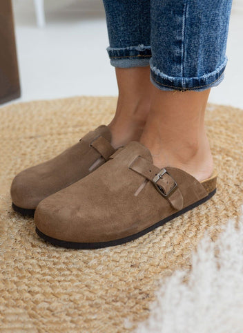 Model foot in taupe suede clog with buckle strap, worn with light blue cuffed jeans, angled view on a woven rug