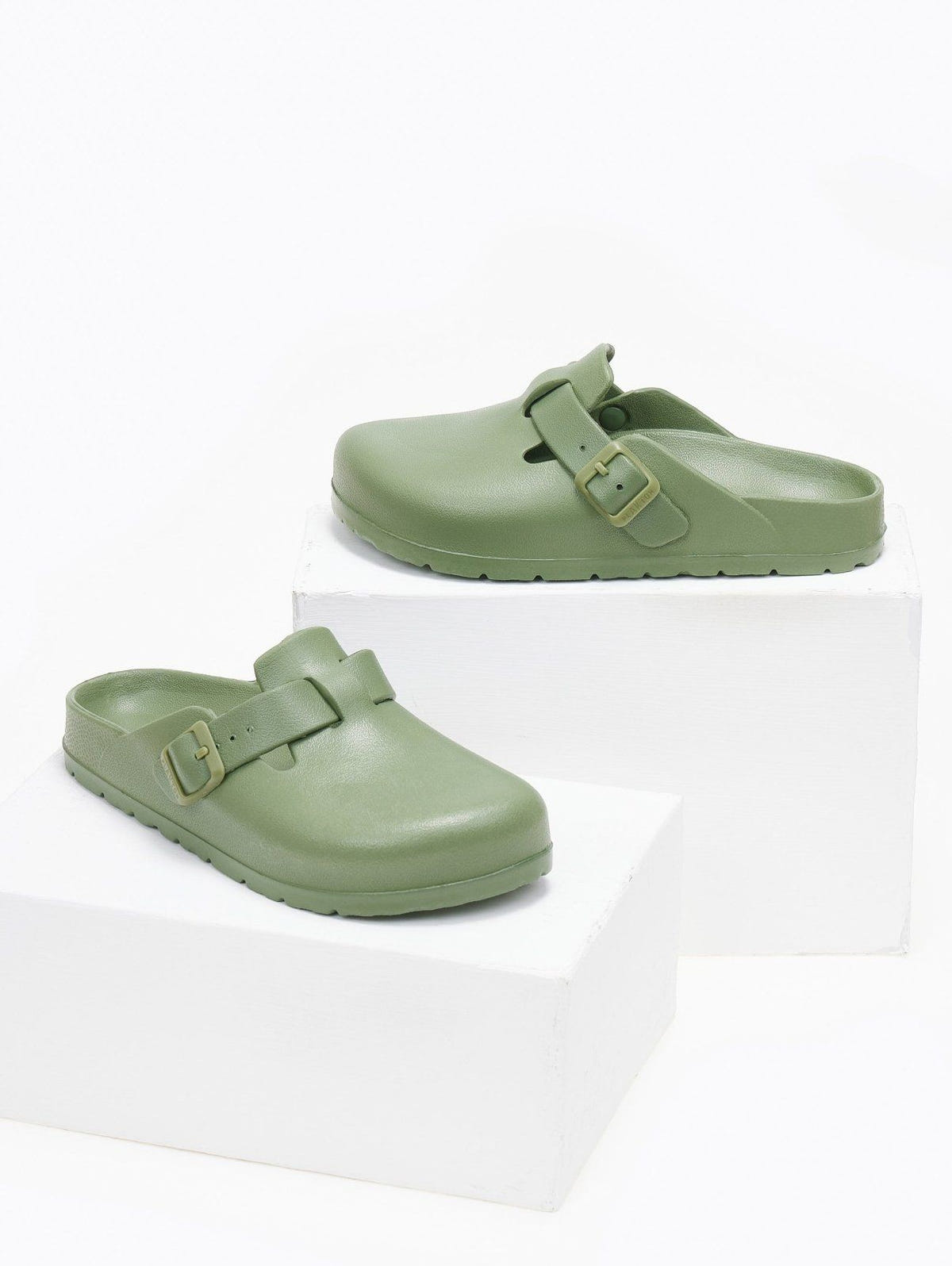 Pair of sage green rubber clogs with buckle straps, angled view on a white display block