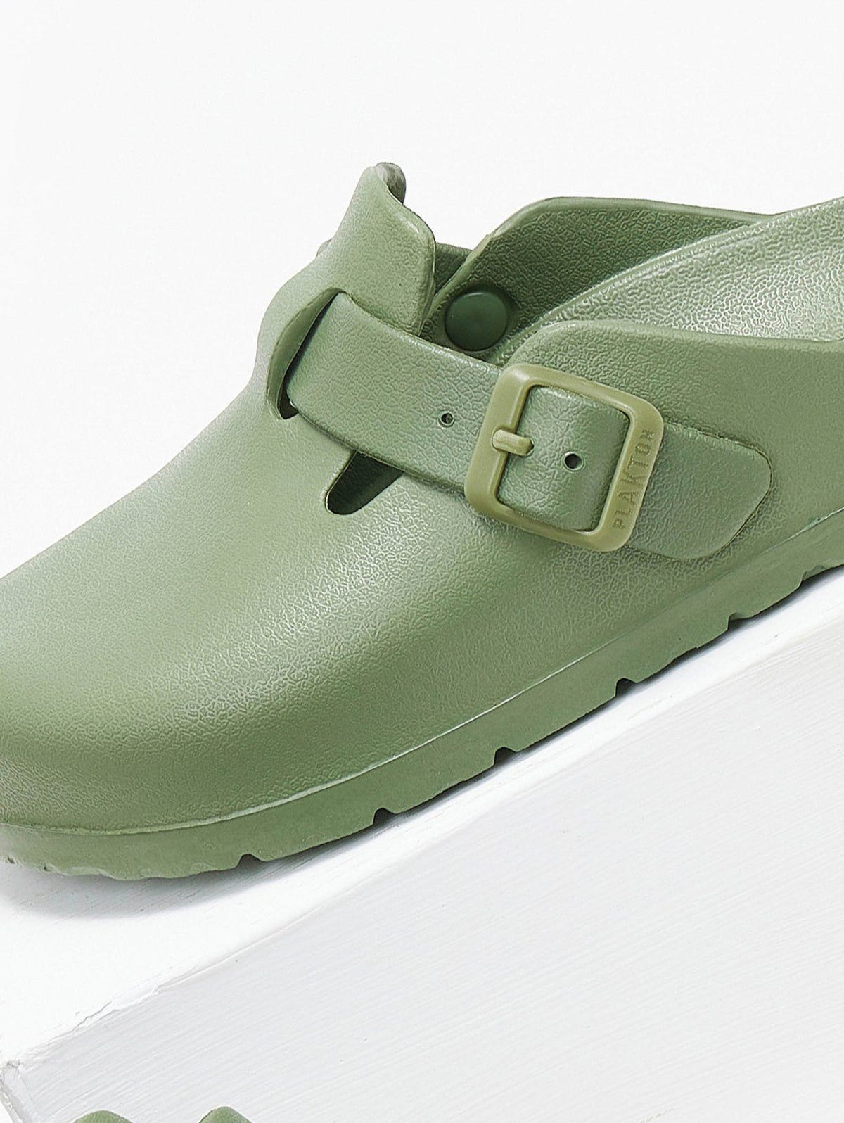 Close-up of sage green rubber clog with buckle strap and sole tread, angled view on a white background