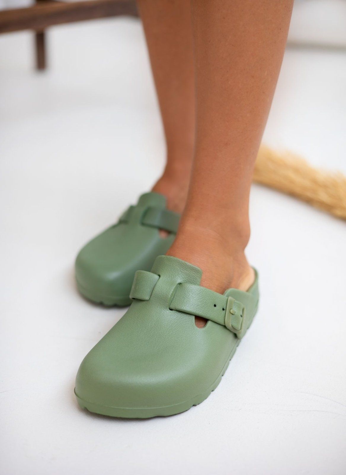 Model feet in sage green rubber clogs with buckle straps, angled view on a light background