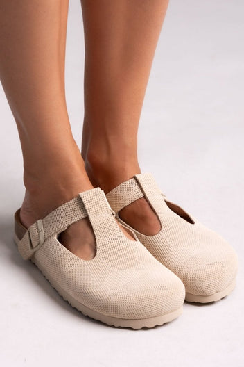 Model feet in cream suede T-strap clogs with cut-out vamp and buckle, angled front view on a light backdrop