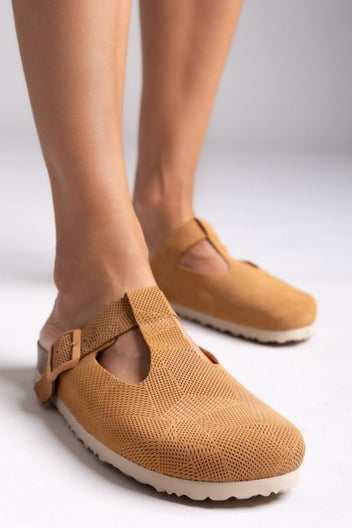 Model legs in tan suede T-strap clogs with cut-out vamp and buckle, angled view on a light grey backdrop