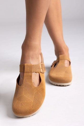 Model legs in tan suede T-strap clogs with cut-out vamp and buckle, front view on a light grey backdrop