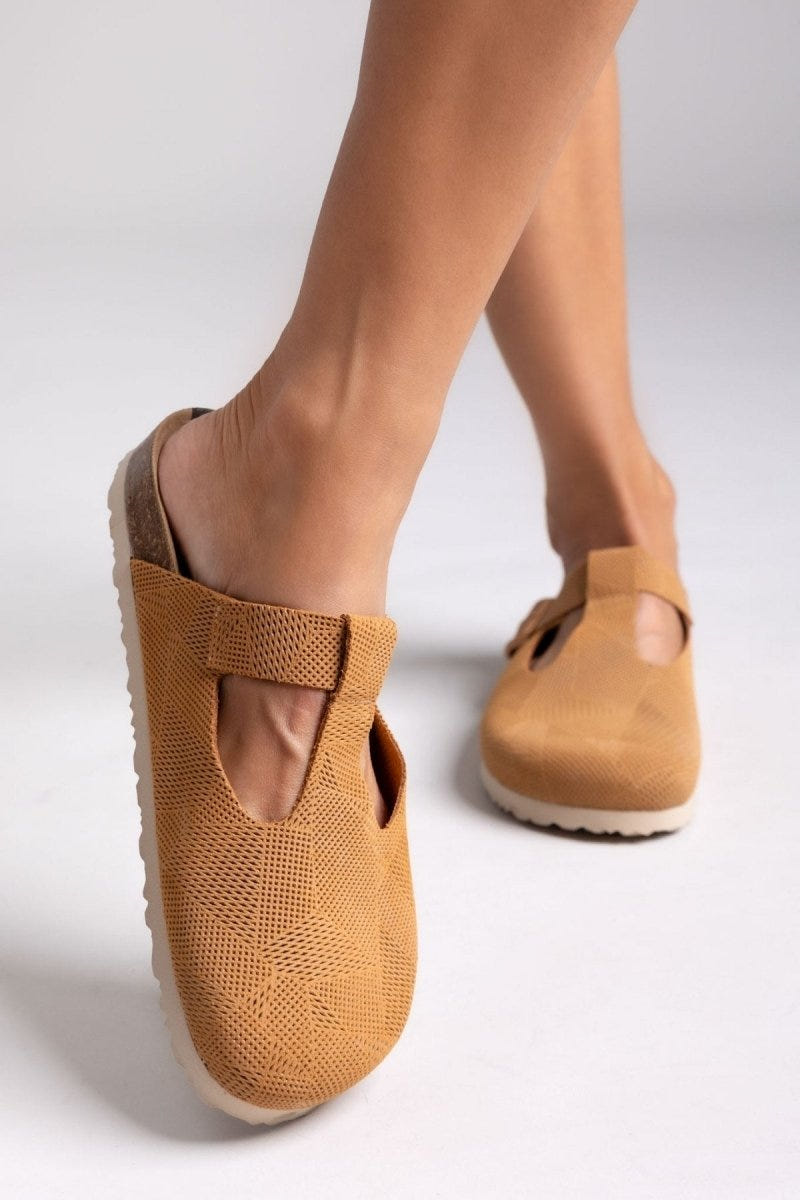 Model leg in tan suede T-strap clog with cut-out vamp and buckle, side view on a light backdrop