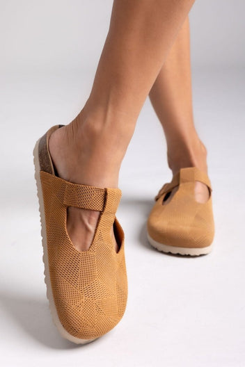 Model leg in tan suede T-strap clog with cut-out vamp and buckle, side view on a light backdrop