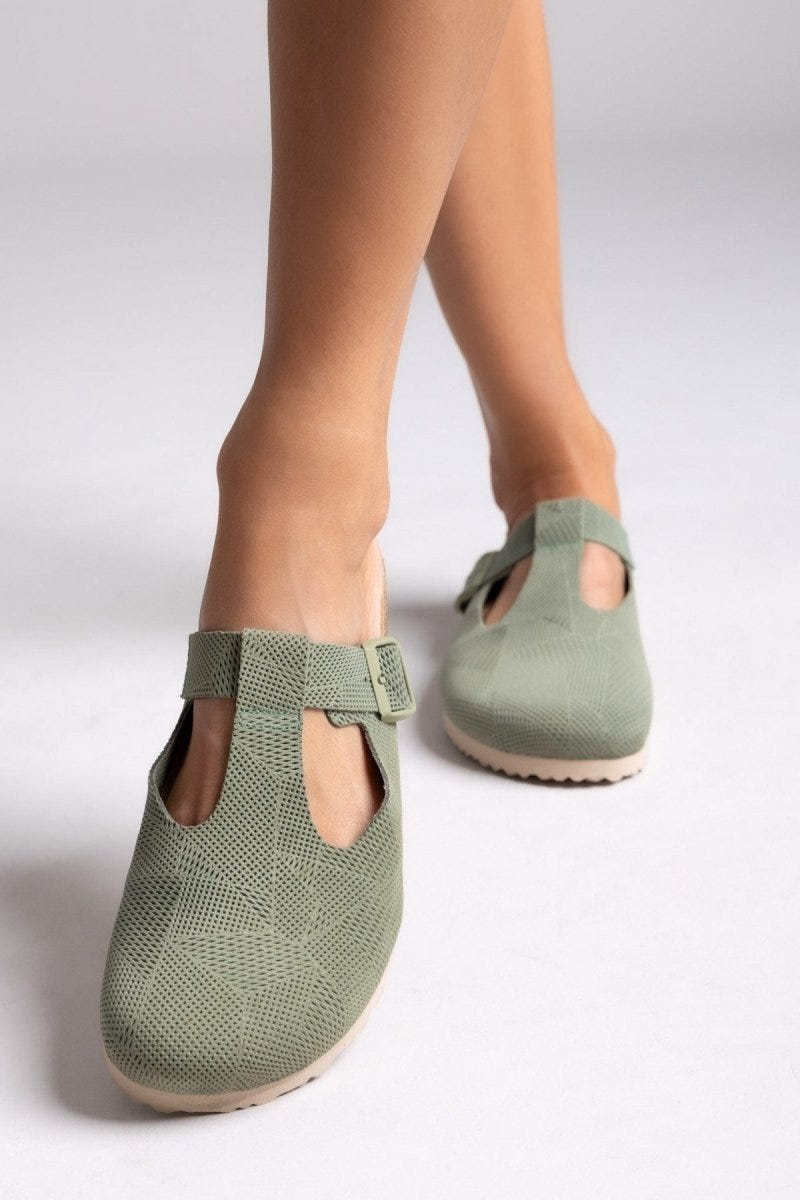 Model legs in sage green suede T-strap clogs with cut-out vamp and buckle, angled view on a light backdrop