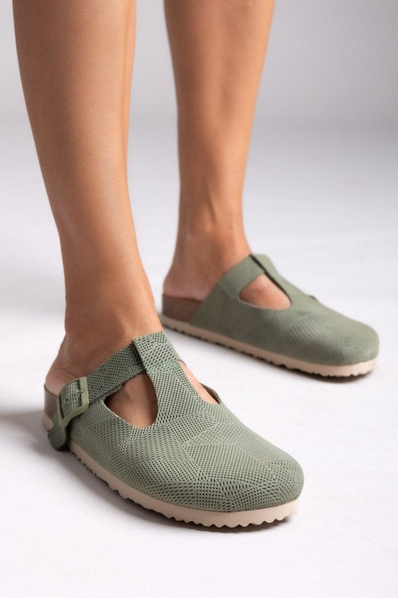 Model legs in sage green suede T-strap clogs with cut-out vamp and buckle, angled view on a light backdrop