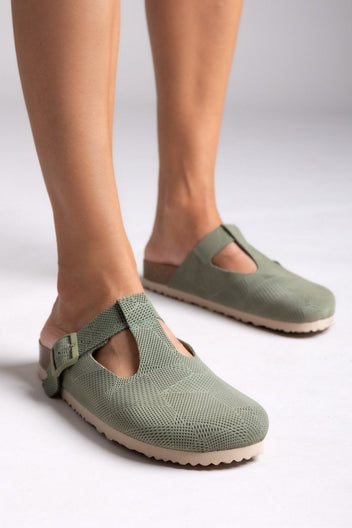 Model legs in sage green suede T-strap clogs with cut-out vamp and buckle, angled view on a light backdrop