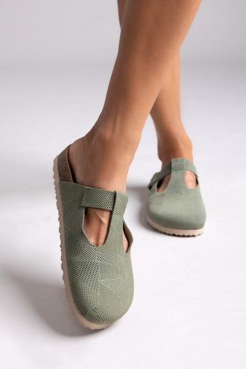 Model leg in sage green suede T-strap clog with cut-out vamp and buckle, angled view on a light backdrop