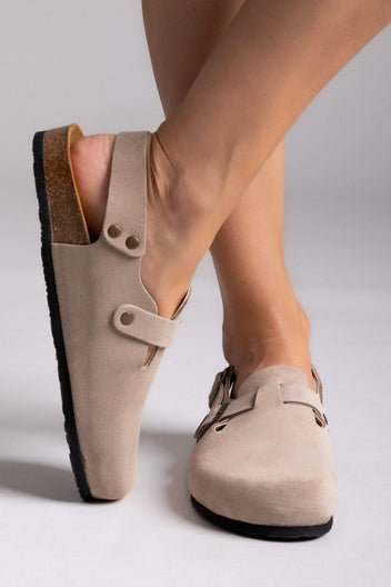 Model leg raised in taupe suede clog with ankle strap and buckle, angled view on a light backdrop