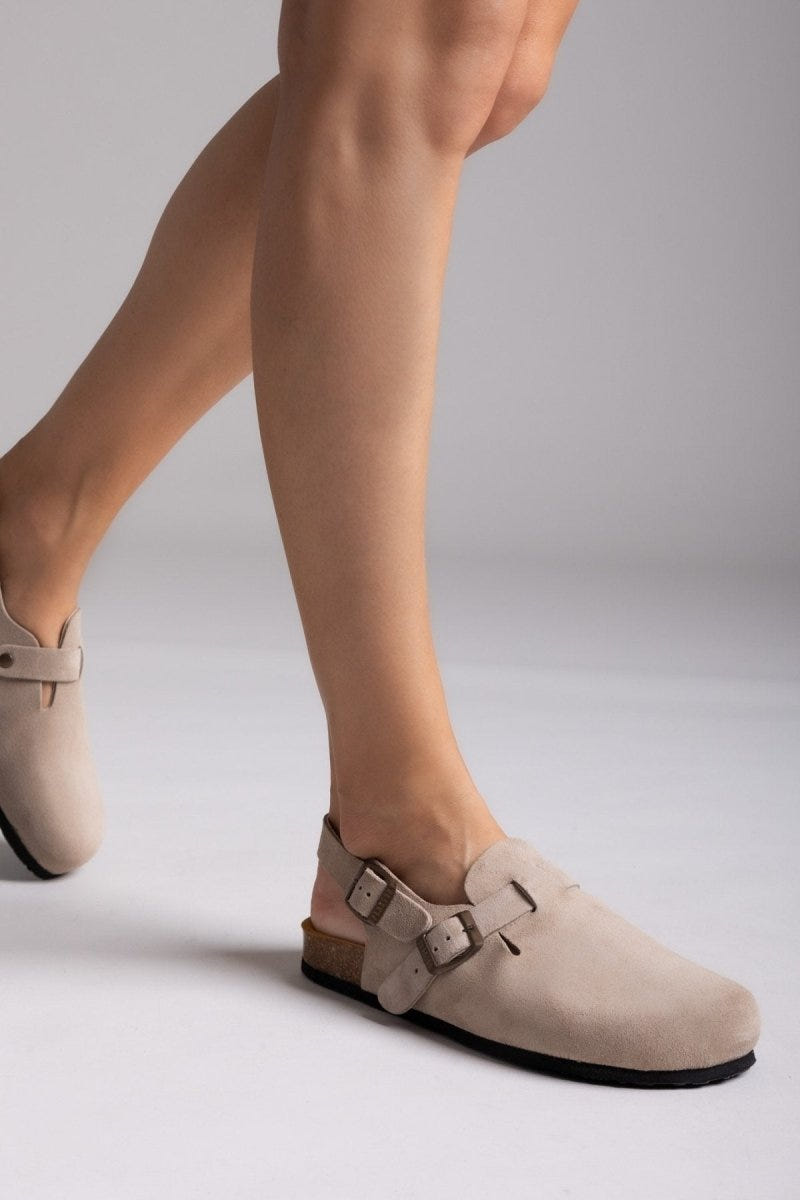 Model leg in taupe suede clog with ankle strap and buckle, angled view on a light backdrop