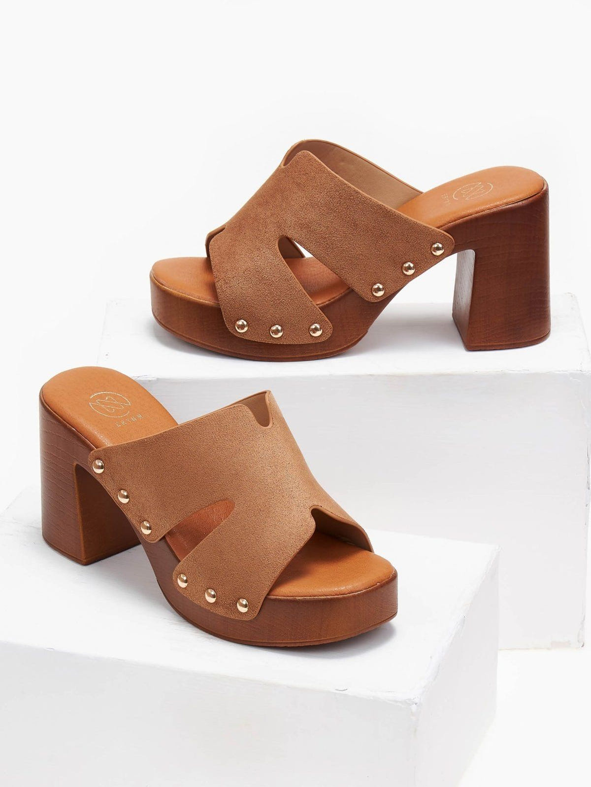 Pair of tan suede block heel slides with cut-out straps, gold studs and wooden-look platform, angled view on a white display block