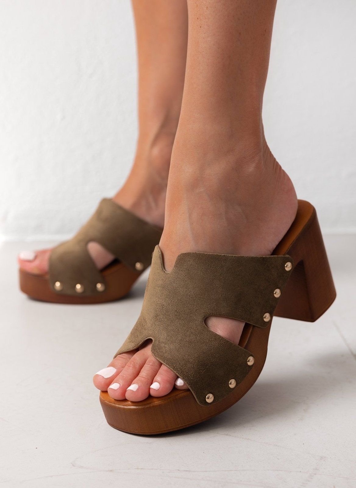 Model feet in olive suede block heel slides with cut-out straps and gold studs, angled view on a light backdrop