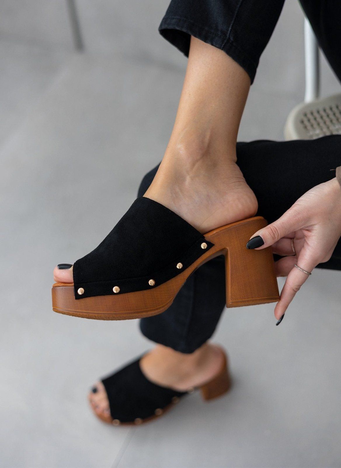 Model feet in black suede block heel slides with gold studs and wooden-look platform, worn with dark trousers, angled view