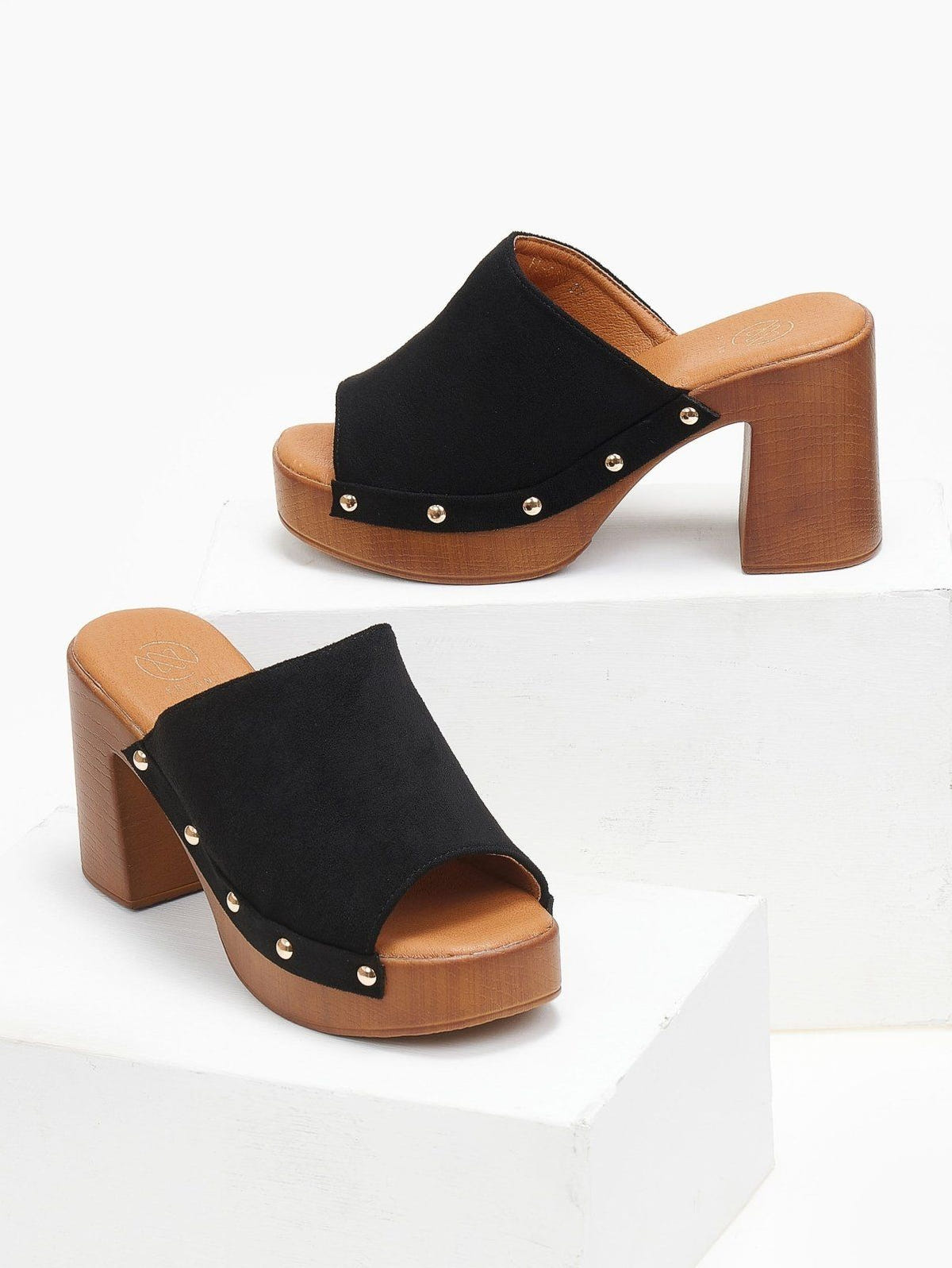 Pair of black suede block heel slides with gold studs and wooden-look platform, angled view on a white display block