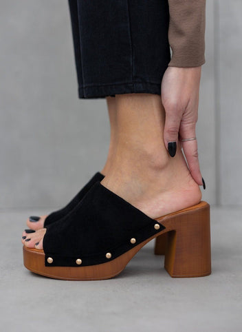 Model foot in black suede block heel slide with gold studs and wooden-look platform, worn with dark trousers, angled view
