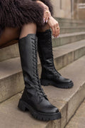 Model seated on stone steps wearing black leather knee-high lace-up boots with chunky lug soles, front view