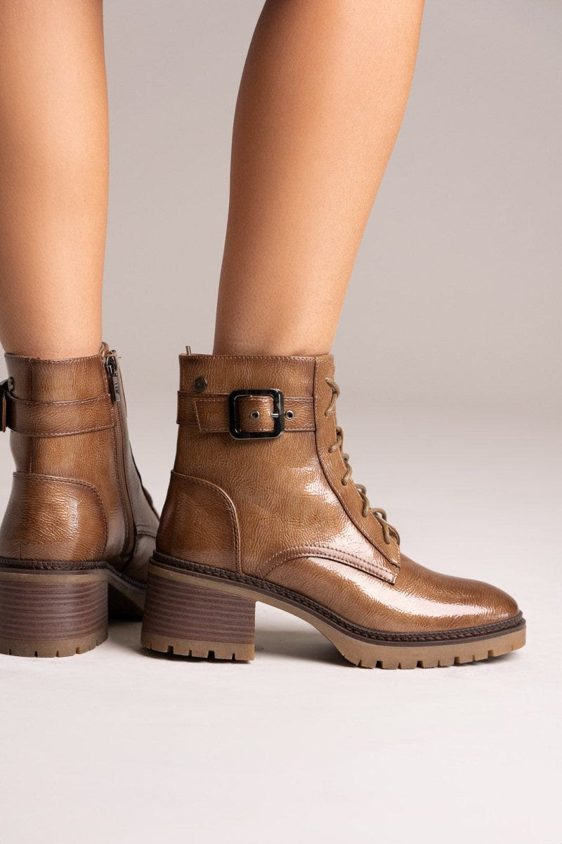 Model legs in brown leather lace-up ankle boots with buckle straps and block heels, angled view on a light backdrop