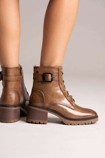 Model legs in brown leather lace-up ankle boots with buckle straps and block heels, angled view on a light backdrop