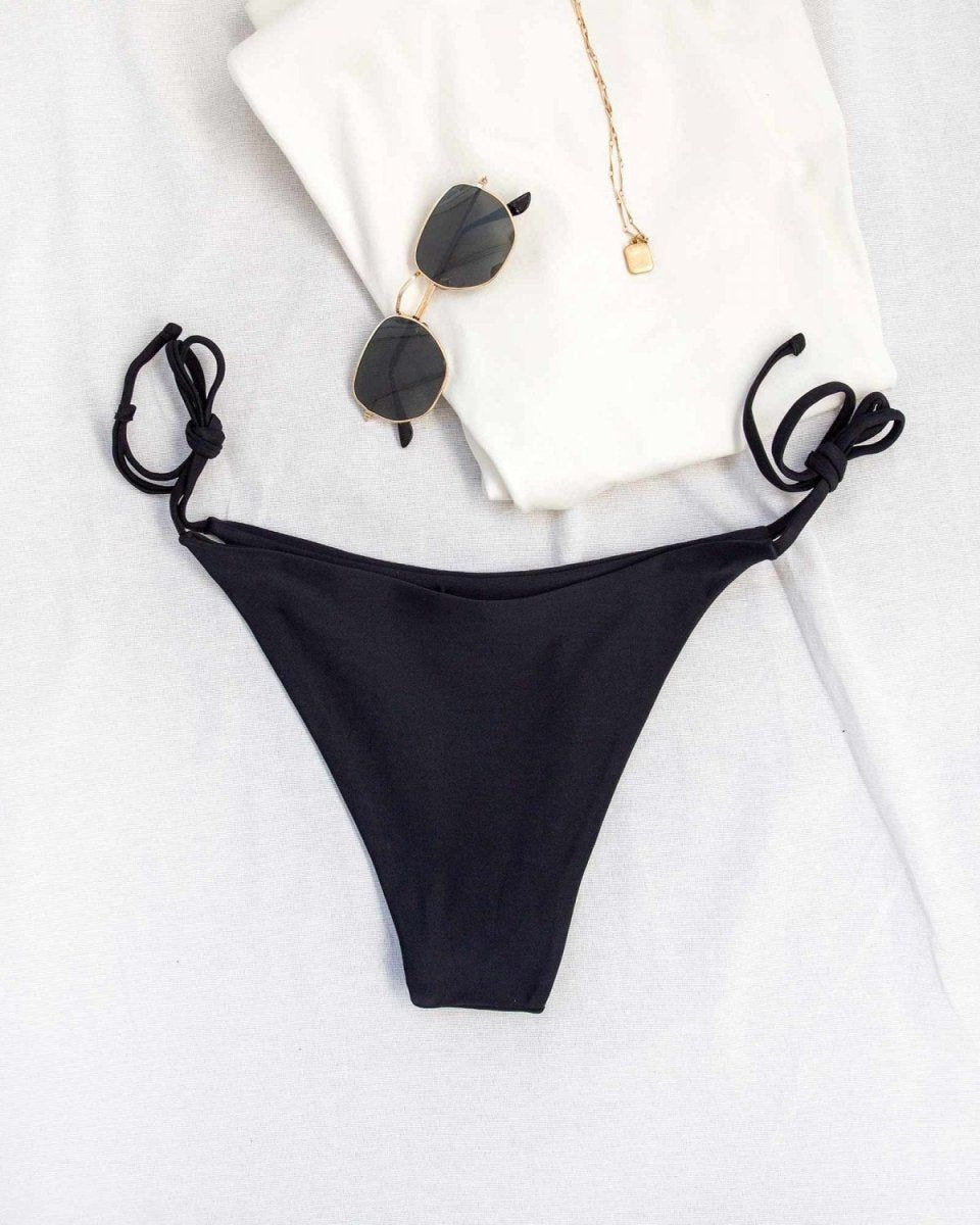 Pair of black side-tie bikini bottoms with white top and sunglasses laid flat on a white background