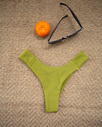 Lime green textured high-cut bikini bottom laid flat beside an orange and sunglasses on a woven beige mat