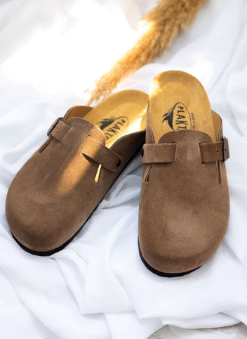 Pair of taupe suede clogs with buckle straps and tan cork footbed on white fabric in top angled view