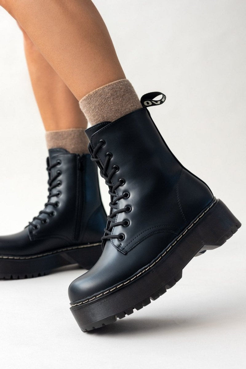 Model wearing black leather lace-up boots with chunky sole and white stitching, side view