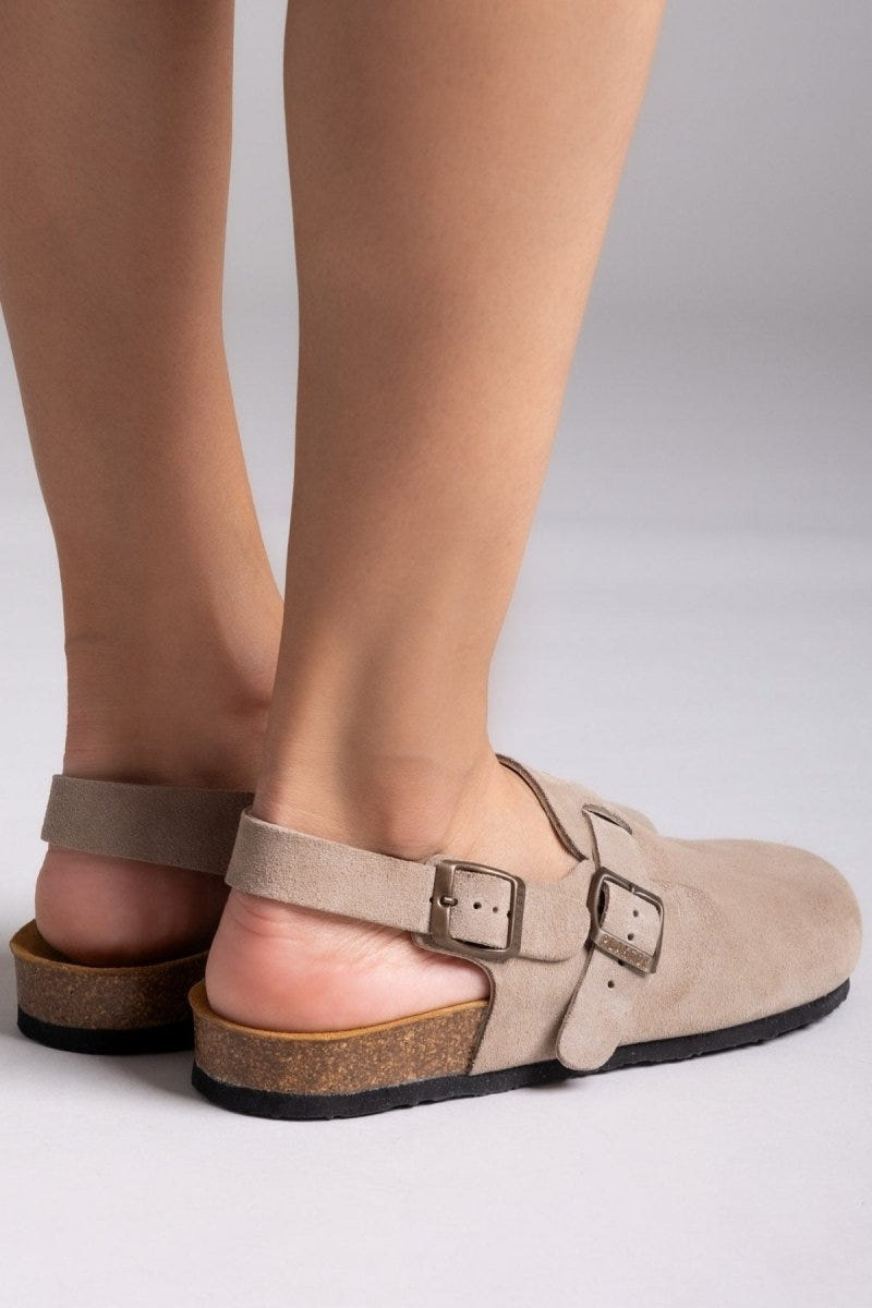 Model feet in taupe suede clogs with ankle strap and buckle, angled view on a light grey backdrop
